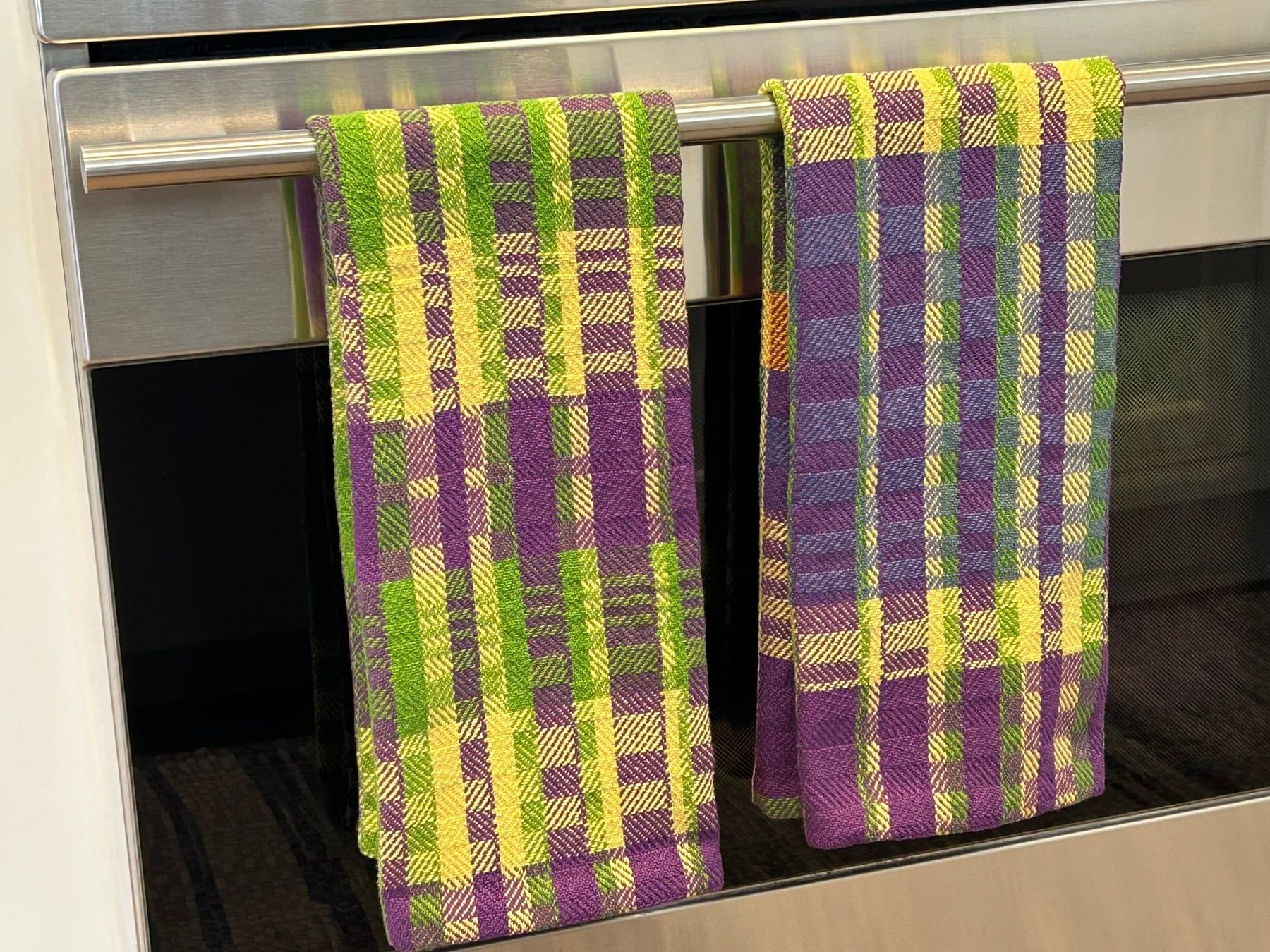 Two plaid tea towels in purple, green and yellow hanging on an oven door